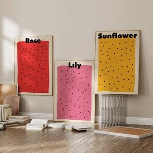 Può includere: Tre stampe incorniciate con disegni astratti di semi neri su uno sfondo colorato. Le stampe sono etichettate "Rosa", "Giglio" e "Girasole" e sono esposte su una parete beige chiaro.