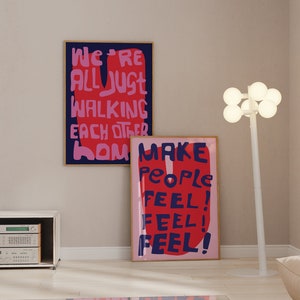 Può includere: Due stampe artistiche incorniciate con testo audace. Una recita "WE'RE ALL JUST WALKING EACH OTHER HOME" in rosa e blu. L'altra dice "MAKE PEOPLE FEEL! FEEL! FEEL!" in rosso e blu. È visibile anche una lampada da terra bianca con luci a globo.