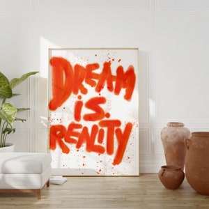 Può includere: Una stampa incorniciata con il testo "DREAM is REALITY" in vernice spray arancione su uno sfondo bianco.
