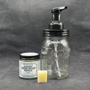 May include: A clear glass mason jar with a black pump dispenser, a small jar of dish soap refills, and a yellow soap cube. The dish soap jar has a white label with black text. The items are for dish soap.