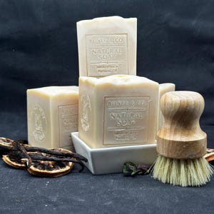 May include: Cream-colored natural soap bars stacked with a wooden brush and dried botanicals. The soap bars are labeled "BILYEU & CO. NATURAL SOAP, handcrafted in Portland, OR." The arrangement includes vanilla beans and dried orange slices.