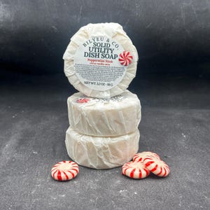 Puede incluir: Dos pastillas redondas de jabón sólido para platos Bilyeu & Co. apiladas y envueltas en papel blanco. La etiqueta superior dice Peppermint Stick, citrus vanilla-mint y net wt. 3.2 oz - 91 g. Tres pequeños caramelos de menta a rayas rojas y blancas.