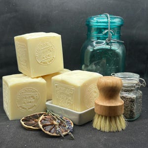 May include: Three pale yellow square soap bars embossed with 100% A-OKI DISH SOAP, a teal glass jar with a wire bail, a small glass jar filled with dried lavender, a wooden scrub brush, and two dried citrus slices with sprigs of lavender.