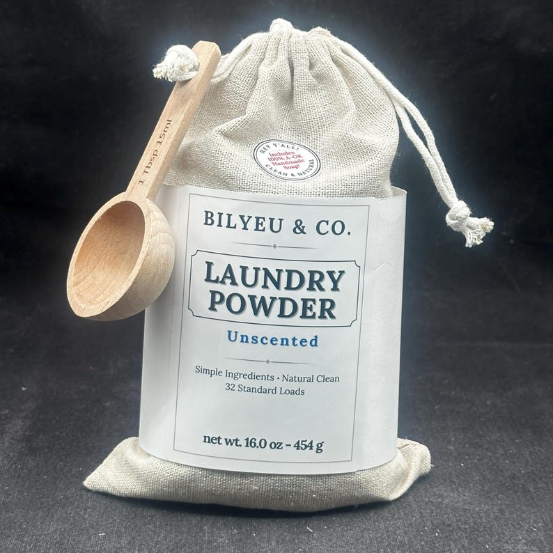 Puede incluir: Una bolsa de tela beige con cord&oacute;n de detergente en polvo Bilyeu & Co. con una etiqueta blanca que dice Laundry Powder Unscented, Simple Ingredients, Natural Clean, 32 Standard Loads, 454 g, y una cuchara de madera marcada con 15ml.