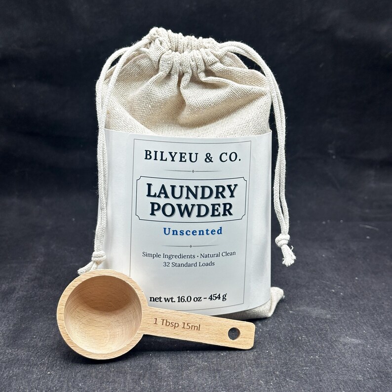 Puede incluir: Una bolsa de tela beige con cord&oacute;n que contiene detergente en polvo con una etiqueta blanca que dice BILYEU & CO. LAUNDRY POWDER Unscented Simple Ingredients Natural Clean 32 Standard Loads net wt. 16.0 oz - 454 g, junto a una cuchara medidora de madera.