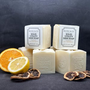 May include: Several blocks of Bilyeu & Co. solid utility dish soap in off-white packaging with black text reading Citrus Zest, lemon, orange & grapefruit, net wt. 11 oz - 312 g. Accompanied by a fresh orange slice, a lemon slice, and dried citrus wheels.