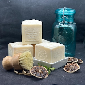 May include: Three square bars of off-white handmade natural soap with embossed text, a wooden scrub brush with light bristles, a teal glass jar with a wire bail, and several dried citrus slices arranged on a dark surface.