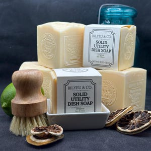 May include: Several blocks of Bilyeu & Co. solid utility dish soap in cream color, each wrapped in a white paper label with black text. A wooden dish brush with light bristles, a fresh lime, and dried lime slices are arranged nearby.