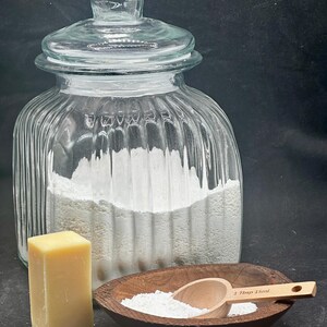 Puede incluir: Un frasco de vidrio transparente acanalado con tapa lleno de polvo blanco, un bloque rectangular de jab&oacute;n amarillo y un cuenco de madera oscura que contiene polvo blanco y una peque&ntilde;a cuchara medidora de madera marcada 1 Tbsp 15ml.