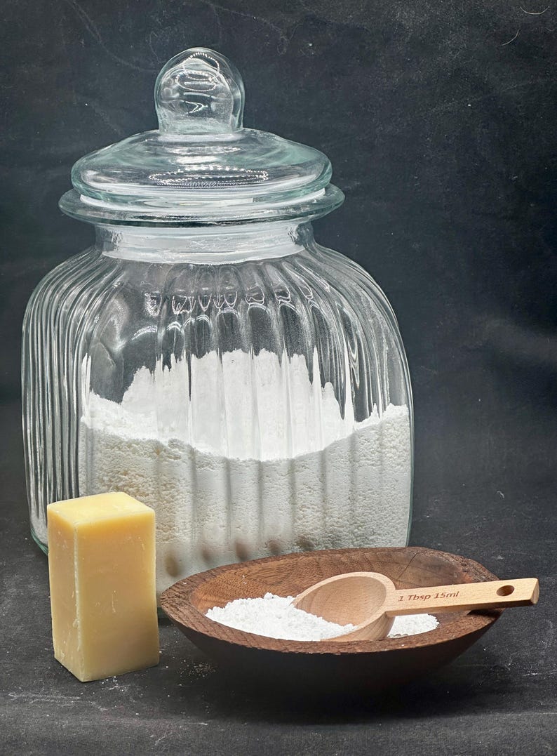 Puede incluir: Un frasco de vidrio transparente acanalado con tapa lleno de polvo blanco, un bloque rectangular de jab&oacute;n amarillo y un cuenco de madera oscura que contiene polvo blanco y una peque&ntilde;a cuchara medidora de madera marcada 1 Tbsp 15ml.