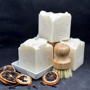 May include: Three square blocks of off-white handmade natural soap with embossed text reading Bilyeu & Co. Handmade Natural Soap. A wooden dish brush with light bristles sits beside the soap, accompanied by dried orange slices and cloves.