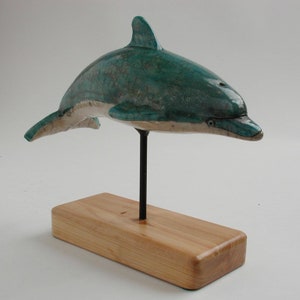 Könnte beinhalten: Eine Delfin-Skulptur aus Keramik mit teal-farbener Glasur und weißen Akzenten. Der Delfin ist auf einem Holzsockel mit einem schwarzen Metallständer montiert.