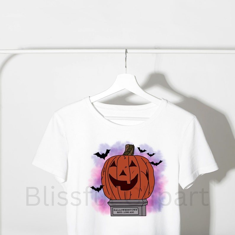 Op de afbeelding: Wit T-shirt met een afbeelding van een uitgeholde pompoen op een voetstuk met de tekst "Halloweentown estd. long ago". De achtergrond is een paars en roze aquareleffect met vleermuizen die rond de pompoen vliegen.