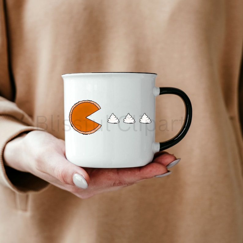 May include: White ceramic mug with a black handle and rim. The mug features a Pac-Man-inspired design with a pie slice and three whipped cream dollops. The mug is held in a person's hands.