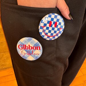 Op de afbeelding: Twee ronde buttons worden getoond. Eén button heeft een blauw-wit geblokt patroon met een rode smiley. De andere button heeft een blauw-wit ruitjespatroon met de rode woorden "Gibbon Buffaloes".