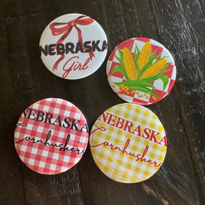 May include: Four round buttons with Nebraska-themed designs. One button has a red bow and the text "NEBRASKA Girl". Another features corn on a red and white checkered background. The other two buttons say "NEBRASKA Cornhuskers" on red and yellow checkered backgrounds.