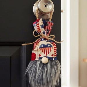 May include: A hanging gnome ornament featuring a fabric hat with a red, white, and blue heart and star pattern. The gnome has a round wooden nose and a long, shaggy gray faux fur beard. It is suspended by a jute twine loop from a metal doorknob.
