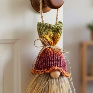 May include: A hanging gnome ornament featuring a knitted hat with a gradient of green, yellow, and deep red yarn. The gnome has a round wooden bead nose and a long, straight, light blonde faux fur beard. A jute twine loop is attached for hanging.