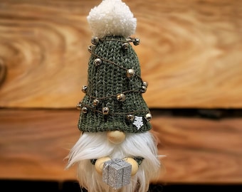 Könnte beinhalten: Ein grüner Häkel-Gnome mit einem weißen Bommelhut mit silbernen Glöckchen und einer weißen Schneeflocke. Der Gnome hält eine silberne Geschenkbox. Der Gnome steht auf einer Holzoberfläche.