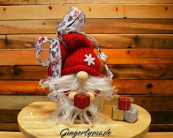 May include: A red and white gnome with a white beard holding a red gift box. The gnome is wearing a red knit hat with a white snowflake on it. The gnome is sitting on a wooden table with a white and gold gift box and a red gift box behind it. The text "Gingerlymade" is written on the table.