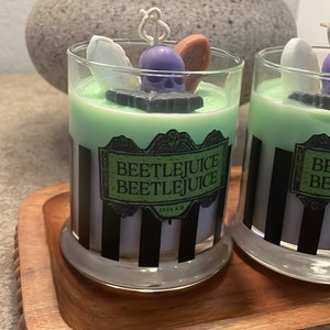 May include: Two green and white striped glass candle jars with black and white labels that say "Beetlejuice Beetlejuice 2021 A.D." The candles are on a wooden tray with a beaded edge.