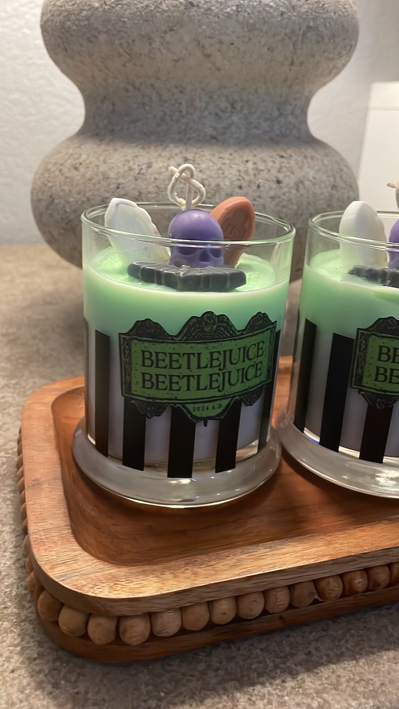 May include: Two green and white striped glass candle jars with black and white labels that say "Beetlejuice Beetlejuice 2021 A.D." The candles are on a wooden tray with a beaded edge.