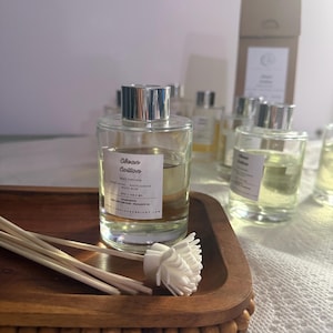May include: A clear glass reed diffuser bottle with a silver cap and a white label reading Clean Cotton, Reed Diffuser, Fresh Ozone, White Jasmine, White Musk, 4oz / 118.2 ml. Several wooden reeds with a white flower-shaped topper rest on a wooden tray.
