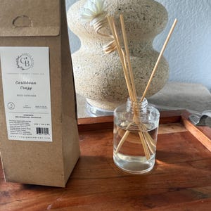 Może przedstawiać: Przezroczysty szklany dyfuzor trzcinowy z drewnianymi trzcinami i małym białym kwiatem. Dyfuzor stoi na drewnianej tacy obok brązowego papierowego opakowania z napisem "Caribbean Crazy" i marką "Candles & Bright".