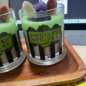 May include: Two glass candle jars with black and white striped designs. The jars are filled with green, white, and brown wax. The jars are decorated with a skull, a tombstone, and a ghost. The jars are labeled "Beetlejuice Beetlejuice 2024 A.D."