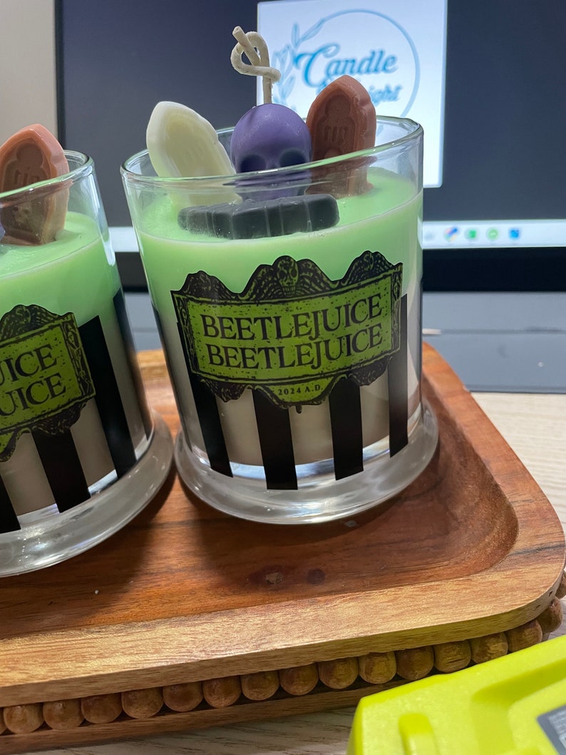 May include: Two glass candle jars with black and white striped designs. The jars are filled with green, white, and brown wax. The jars are decorated with a skull, a tombstone, and a ghost. The jars are labeled "Beetlejuice Beetlejuice 2024 A.D."