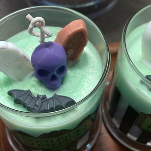 May include: Two green scented candles with Halloween-themed decorations. The candles are in glass jars with black and white striped labels that say "Beetlejuice Beetlejuice Beetlejuice". The decorations include a purple skull, a white tombstone, a brown coffin, and a black bat.