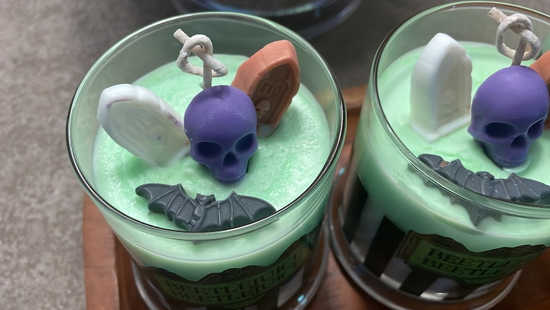 May include: Two green scented candles with Halloween-themed decorations. The candles are in glass jars with black and white striped labels that say "Beetlejuice Beetlejuice Beetlejuice". The decorations include a purple skull, a white tombstone, a brown coffin, and a black bat.