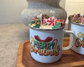 May include: White enamel mug with a "Merry Christmas" graphic, filled with decorative candies and small Christmas tree and gift-shaped decorations. The mug sits on a wooden tray with a beaded edge, perfect for holiday gifting.