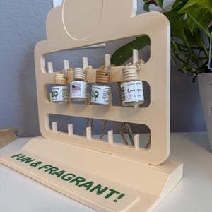 Puede incluir: Un expositor de madera de color claro con el texto FUN & FRAGRANT! en letras verdes. Cuatro frascos de vidrio pequeños con tapones de madera y cordones para colgar están suspendidos del soporte. Cada frasco tiene una etiqueta blanca.