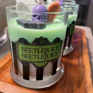 May include: Three green and white striped glass candle jars with black and white labels that say "Beetlejuice Beetlejuice" and "2024 A.D." The candles are topped with a green wax layer and decorated with purple, white, and orange wax figures.