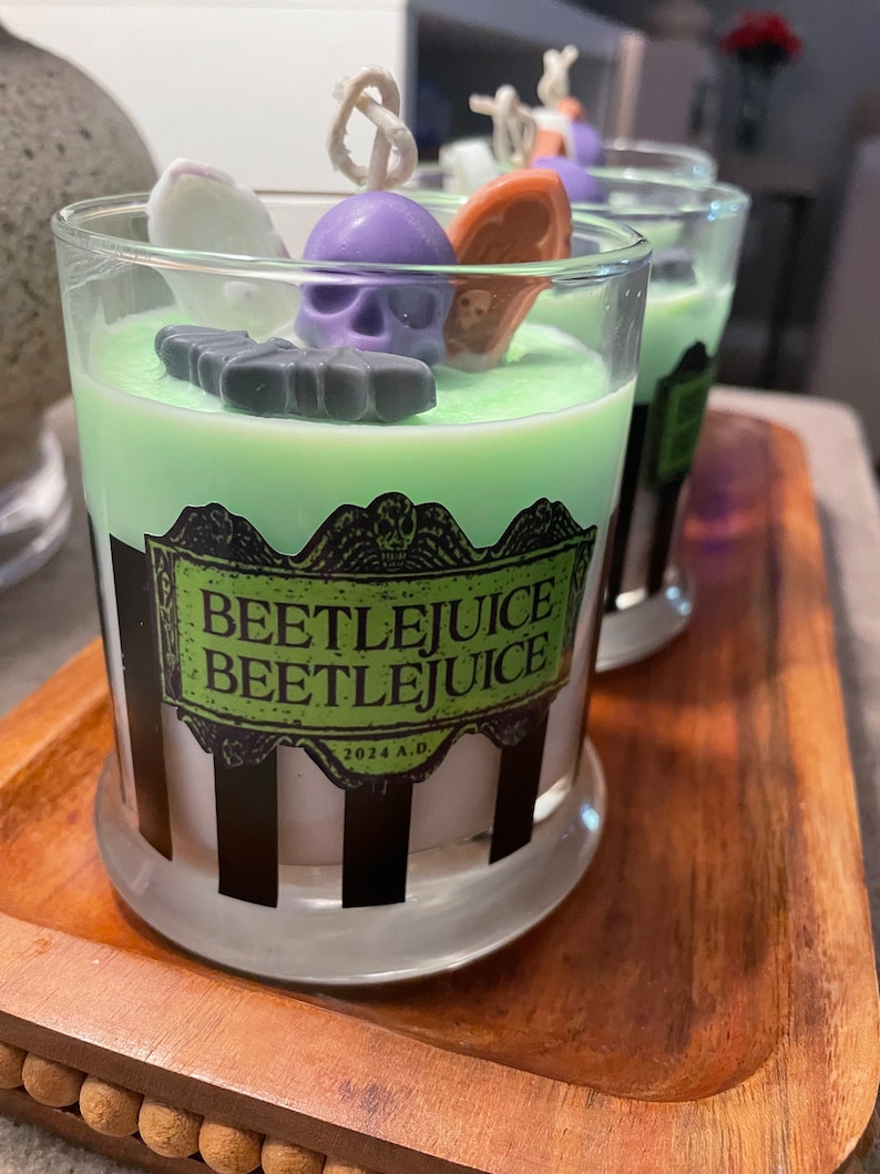 May include: Three green and white striped glass candle jars with black and white labels that say "Beetlejuice Beetlejuice" and "2024 A.D." The candles are topped with a green wax layer and decorated with purple, white, and orange wax figures.
