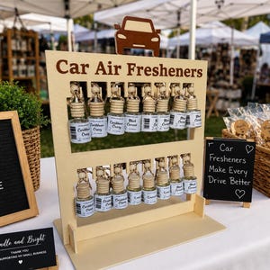 May include: A light-colored wooden display stand with a car icon at the top and the text Car Air Fresheners. It holds two rows of small glass bottles with wooden caps and hanging cords, each labeled with scents like Caribbean Crazy and Black Currant.
