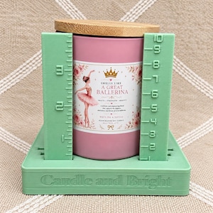 May include: A pink candle in a cylindrical container with a bamboo lid, placed on a light green stand with integrated rulers showing a height of 10 inches. The label features a ballerina illustration and text reading Smells Like A Great Ballerina.