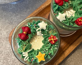 Puede incluir: Dos velas de vidrio transparente con un diseño de corona navideña. Las coronas están hechas de glaseado verde y decoradas con lazos rojos, adornos en forma de estrella y de regalo, y chispas doradas. Las velas están sobre una bandeja de madera.
