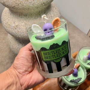 May include: A green and white striped candle jar with a black and white label that reads "Beetlejuice Beetlejuice 1972 A.D.". The candle jar is decorated with a purple skull, a black bat, a white coffin, and a brown tombstone.