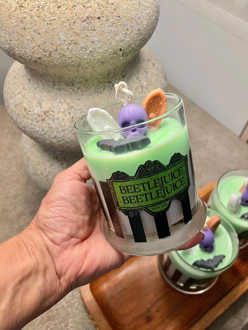 May include: A green and white striped candle jar with a black and white label that reads "Beetlejuice Beetlejuice 1972 A.D.". The candle jar is decorated with a purple skull, a black bat, a white coffin, and a brown tombstone.