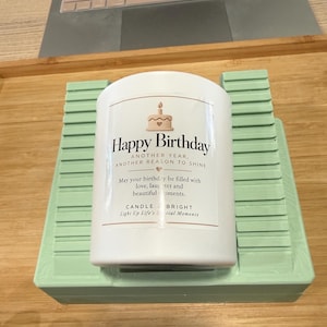 May include: A white cylindrical candle with a label featuring a cake illustration and the text Happy Birthday, Another Year, Another Reason To Shine, May your birthday be filled with love, laughter and beautiful moments, Candle Bright.