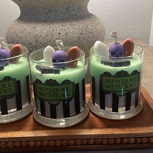 May include: Three glass candle jars with green and black striped labels that read "Beetlejuice Beetlejuice". Each jar has a green wax base with a purple skull, white ghost, and orange tombstone on top.