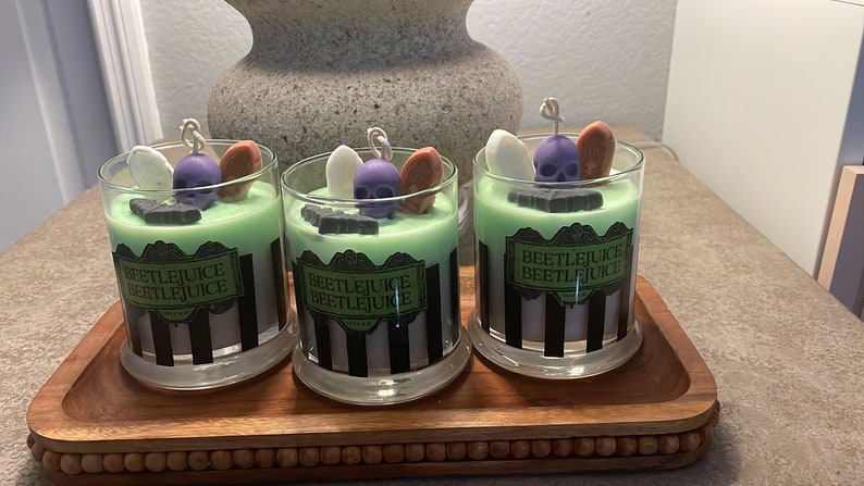 May include: Three glass candle jars with green and black striped labels that read "Beetlejuice Beetlejuice". Each jar has a green wax base with a purple skull, white ghost, and orange tombstone on top.