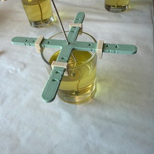 May include: A light green plastic candle wick centring tool with four adjustable arms resting on a clear glass jar filled with yellow liquid wax. The tool features a central hole for the wick and small clips to secure the position.