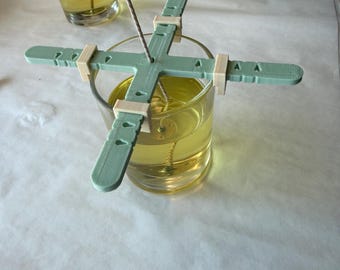 May include: A light green plastic candle wick centering tool with four adjustable arms resting on a clear glass jar filled with yellow liquid wax. The tool features a central hole for the wick and small clips to secure the position.