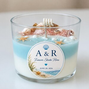 May include: A clear glass candle with a layered blue and white wax base, topped with embedded seashells and starfish. A circular label features a blue seashell icon, the text A & R, Forever Starts Here, a blue heart, and JUNE 20, 2026.