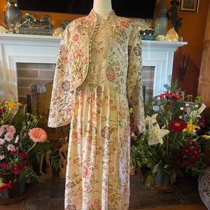 May include: A vintage-style cream-colored dress and jacket set. The dress features a floral pattern in pink, brown, and green. The matching jacket is quilted with a similar floral design and has a button-up front. The outfit is displayed on a mannequin.