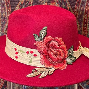 May include: A red felt fedora hat with a beige ribbon and a large embroidered rose in red, orange, and green.