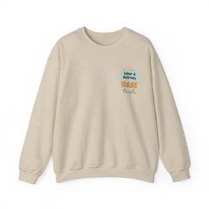 Op de afbeelding: Een beige sweatshirt met een afbeelding van een cupcake met de tekst "labor & delivery tech" op de voorkant.